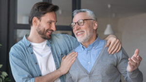 A younger man with dark hair and a beard smiles and places his arm around an older man with glasses, gray hair, and a beard. The older man is also smiling, and they both appear to be enjoying each other's company. They stand indoors, dressed casually.