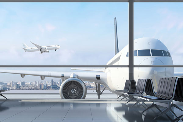 A large white airplane is parked at a gate at the airport, viewed through the floor-to-ceiling windows of a terminal waiting area with empty black chairs. Another airplane is taking off in the background, with a city skyline visible in the distance under a clear blue sky.