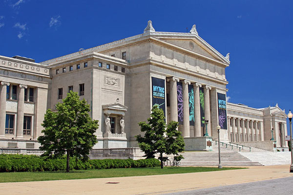 A large neoclassical building with tall columns and banners hanging on its front, alongside statues and neatly trimmed shrubs. Steps lead up to the entrance, framed by clear blue skies.