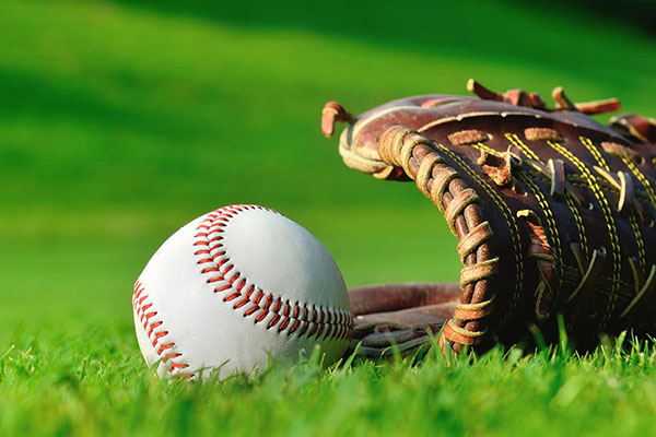 A close-up image of a white baseball with red stitching lying on bright green grass, next to an open brown leather baseball glove. The background is a slightly blurred grassy field.