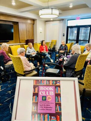 A group of elderly individuals are seated in a circle in a room, engaged in a book club discussion. A sign in the foreground reads, "BOOK CLUB with Isabel Prairie Library April 10, 2024, 1 PM, Wednesday, Cadillac Theater," placed on an easel.