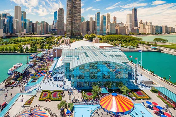 A vibrant aerial view of Navy Pier in Chicago, featuring an amusement park with a carousel in the foreground, a large glass-covered building at the center, and the city's skyline with numerous high-rise buildings and Lake Michigan in the background under a partly cloudy sky.