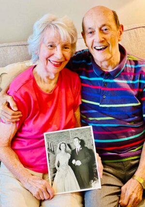 An elderly couple sits on a couch, smiling and embracing each other. The woman, wearing a pink shirt, holds a black-and-white wedding photo. The man, in a striped shirt, sits next to her with his arm around her shoulders. Both appear happy and joyful.