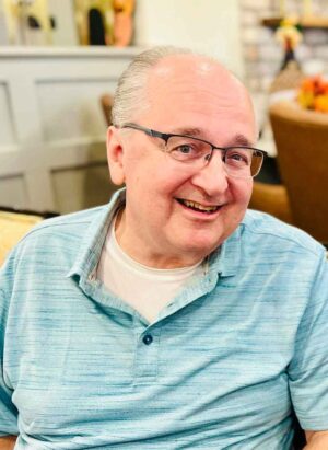 A smiling older man with glasses and a receding hairline is wearing a light blue polo shirt. He is sitting indoors on a chair with a cozy background, including blurred-out decor elements like flowers and a brick wall.