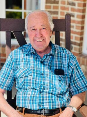 An elderly man with white hair and a friendly smile sits on a wooden chair. He is wearing a blue plaid shirt with a black phone in his chest pocket and brown pants. The background shows a brick wall and a window.