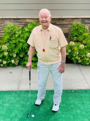 An elderly man, smiling and wearing a light-colored short-sleeve shirt and white pants, stands on a patch of green turf holding a golf putter. He has a red whistle on a lanyard around his neck and a pen in his shirt pocket. Shrubs and a stone wall are in the background.