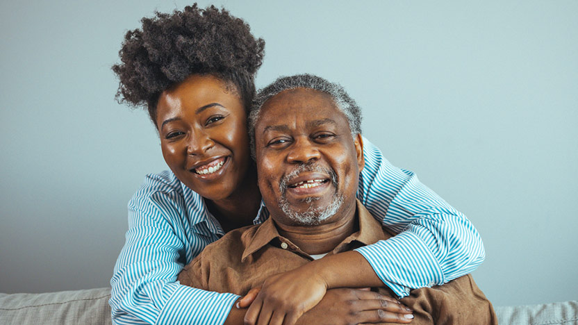 A young woman with curly hair and a striped shirt hugs an older man with gray hair and a brown shirt.