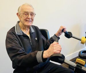 An elderly man with glasses and a smile is seated on a fitness machine, possibly an exercise bike, holding the handlebars. He is wearing a dark jacket with a gray collar and gray cuffs. The background is a plain, light-colored wall.