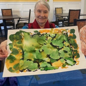 An elderly woman with gray hair and a red scarf smiles as she holds up a colorful abstract painting with green and yellow hues. She is seated at a table in a communal room with white paneled walls and several chairs and tables in the background.