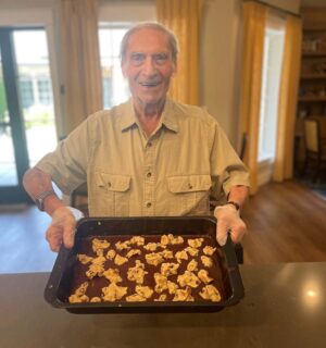 An elderly man with gray hair and wearing a beige shirt stands in a kitchen, smiling and holding a baking tray filled with chocolate and cookie dough. He is wearing white gloves. Yellow curtains cover the windows in the background.