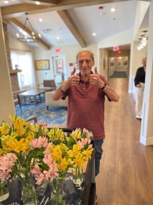 An elderly man wearing a maroon polo shirt and blue jeans gives a double thumbs up while smiling. He stands in a brightly lit room with wooden floors, where a table adorned with yellow and pink flowers is in the foreground.