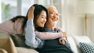 A young woman warmly hugs an older man while they sit on a couch, both smiling joyfully.