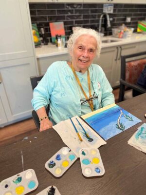 An elderly person with curly white hair and a light blue shirt is smiling while seated at a table, engaging in a painting activity. They are surrounded by art supplies, including paint palettes and brushes, and a partially completed painting of a tree on a canvas.