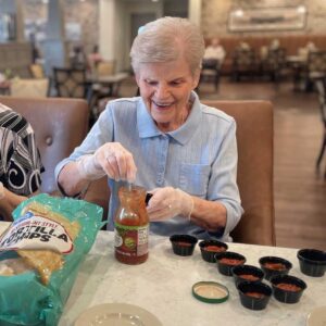 An elderly woman with short grey hair and a light blue blouse smiles while spooning salsa from a jar into small black cups. She sits at a table with tortilla chips and a beige plate beside her. The background shows a well-lit room with other people seated at tables.