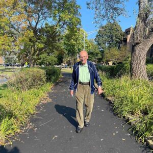 An elderly man walks along a paved pathway in a park on a sunny day. He wears a green shirt, dark blue jacket, and brown pants. Trees with green and autumn-colored leaves line the path, and there are bushes and other greenery around.